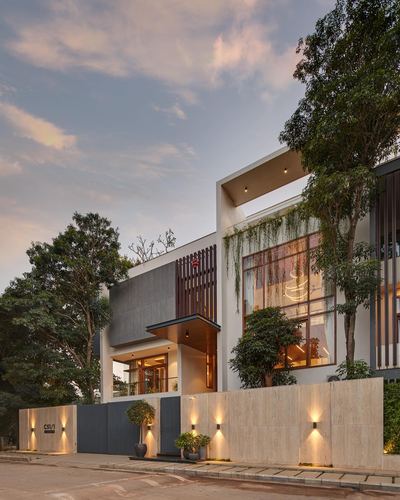 Contemporary Bangalore house facade at dusk with stone boundary wall, wood slats, and lit landscape lighting.