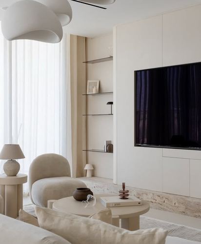 Cream-toned living room with sheer curtains, glass shelving, and a wall-mounted tv above a stone base.