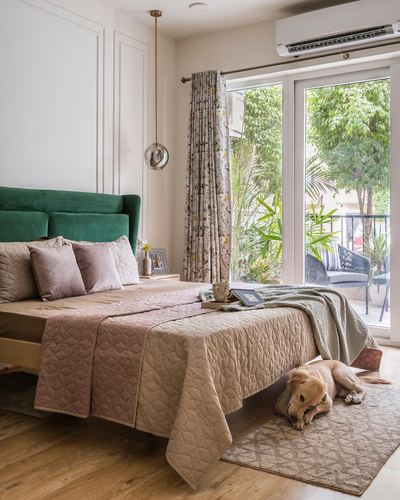 Bedroom with green velvet headboard, pink quilt, floral curtains and dog on rug near glass balcony door