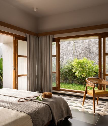 Bedroom corner with wooden windows, cane chair, and garden view through stone wall