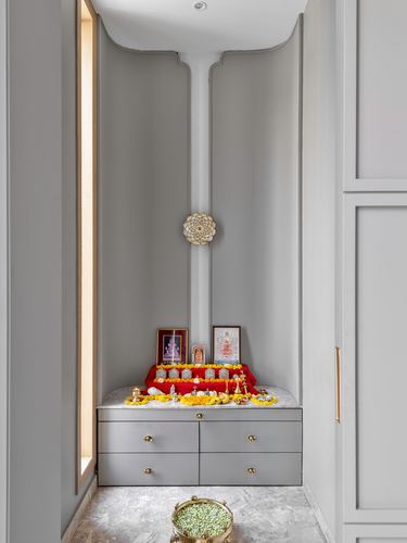 Grey pooja niche with framed deities, marigold flowers, and drawer storage below