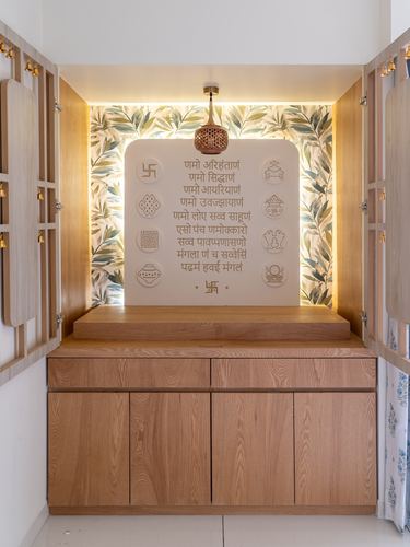 Pooja unit with carved mantra panel, botanical wallpaper backdrop, hanging brass lamp, and wooden cabinetry with bell-hung doors
