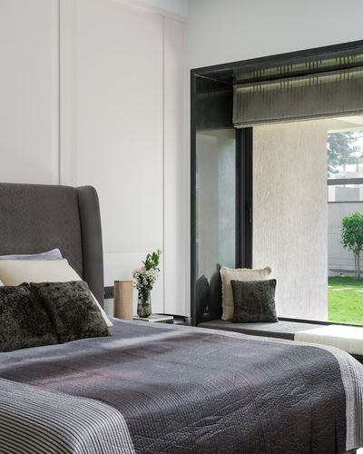 Grey upholstered bed beside a black-framed window seat overlooking a garden