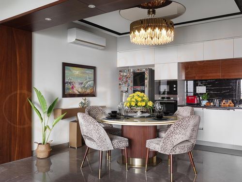 Round dining table with patterned chairs under a tiered chandelier, open to a white kitchen