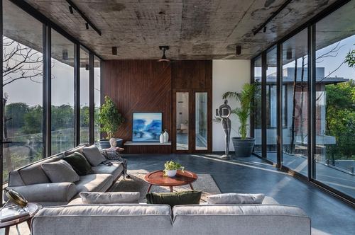 Glass-walled living room with wood-panel accent wall, grey sectional sofa, and greenery views on all sides