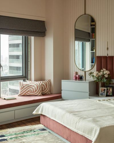 Pink-toned bedroom with window seat, fluted paneling, and oval brass mirror overlooking city skyline