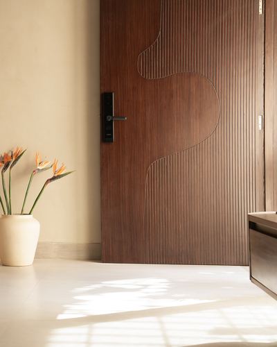 Fluted wood entry door with curved inlay, cream vase with orange flowers beside it, sunlit floor
