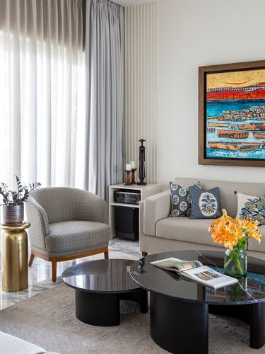 Living room corner with beige sofa, cane armchair, black round coffee tables and colorful framed painting