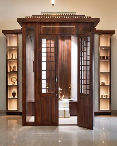 Ornate wooden double doors of a pooja room flanked by lit display shelves with brass idols