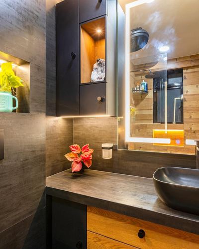 Dark stone bathroom vanity with black vessel sink and backlit mirror reflecting wood-clad shower