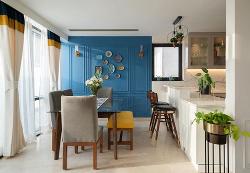 Dining area with glass table, mustard chair, and blue paneled wall hung with decorative plates