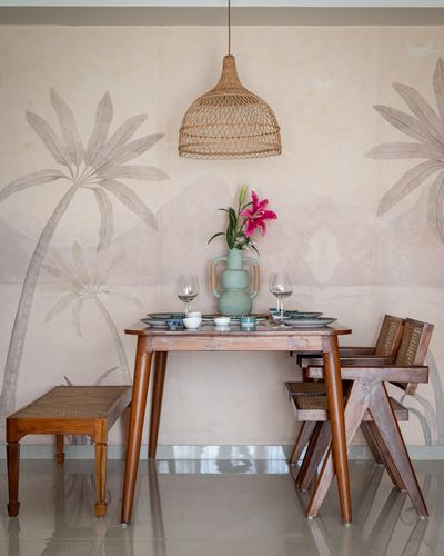 Wood dining table with cane chairs beneath a woven pendant light against a palm-mural wall