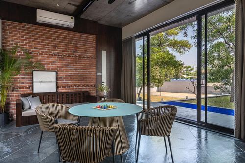 Dining nook with round table and woven chairs beside brick wall, glass doors open to pool and trees.