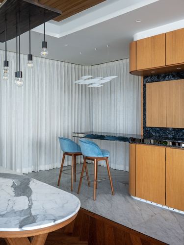 Kitchen counter with blue velvet stools, curved wood cabinets, marble backsplash and sheer curtains