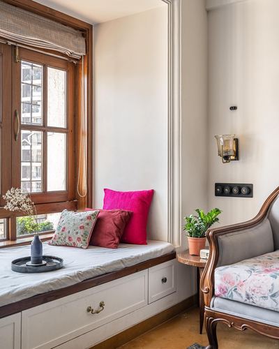 Wooden window seat with pink cushions beside a carved armchair and small plant table