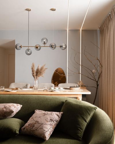 Curved olive-green sofa with floral cushions in foreground, dining table and glass globe chandelier beyond