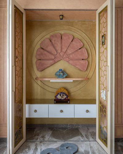 Pooja alcove with fan-shaped pink panel, gold walls, floating white cabinet and patterned glass doors