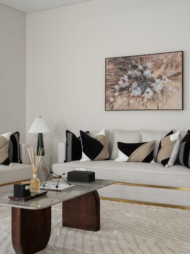 Beige sofa with black and tan geometric pillows beneath a floral painting, marble coffee table in front