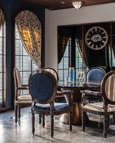 Round marble dining table with blue velvet chairs beneath a large decorative clock and arched draped windows