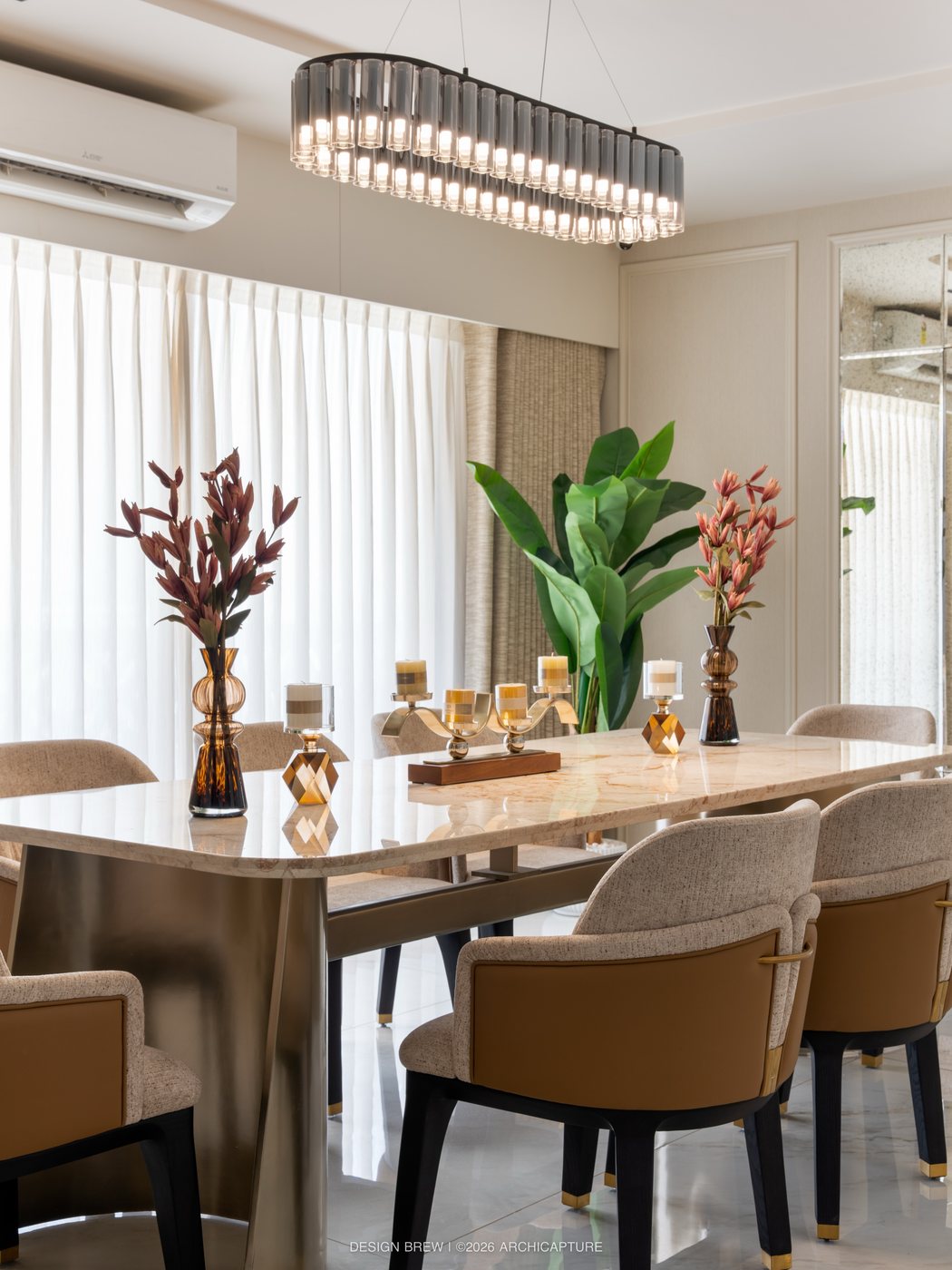 Formal dining table with tan leather chairs beneath a linear tube chandelier and sheer curtained window