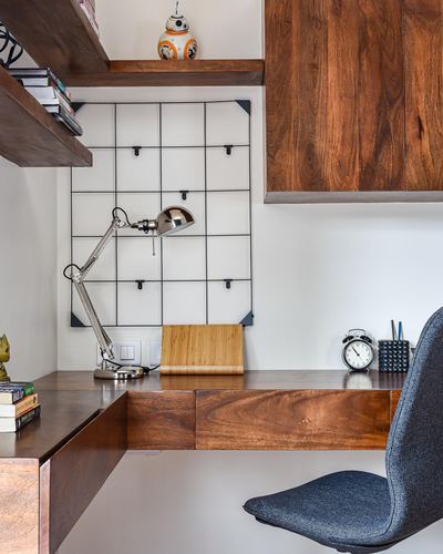 Corner home office with wooden L-shaped desk, chrome lamp, wire memo board, and upper wood cabinet