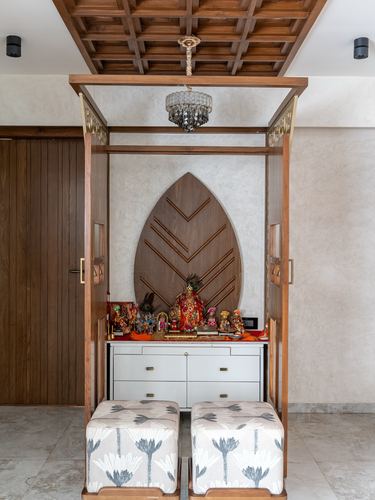 Wooden pooja unit with carved leaf motif backdrop, brass hardware, chandelier, and two floral-print stools in front.