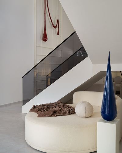Round cream boucle daybed beneath a staircase with glass railing, tall blue ceramic sculpture beside it.