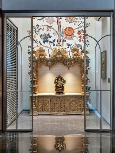 Carved wooden mandir with brass Ganesha idol framed by glass doors and floral mural wall