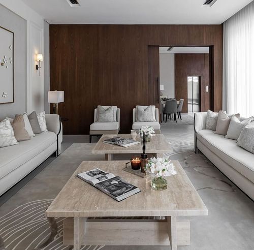 Symmetrical living room with beige sofas, travertine coffee tables, and dark wood paneled wall opening to dining area