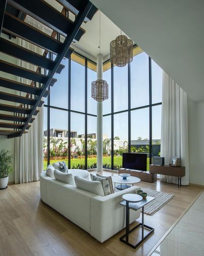 Double-height living room with floating staircase, tall glass wall overlooking garden, white sofa and woven pendant lights