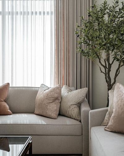 Beige sofa with pink and textured cushions beside sheer curtains and a potted olive tree