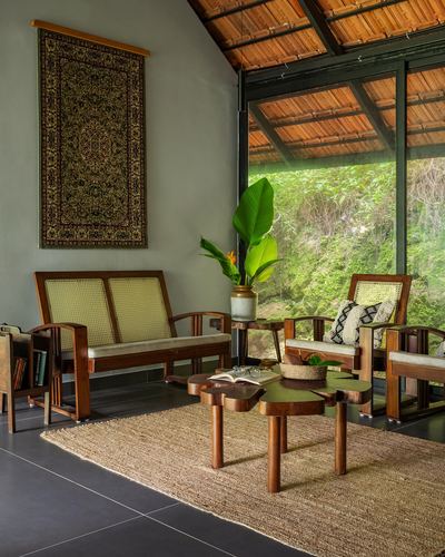 Cane-backed wooden seating and carved coffee table facing glass wall overlooking forest, under exposed wood-slat roof