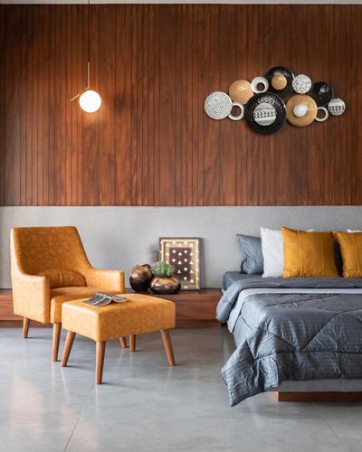 Bedroom with wood-paneled wall, mustard armchair and ottoman, and grey bed with mustard cushions