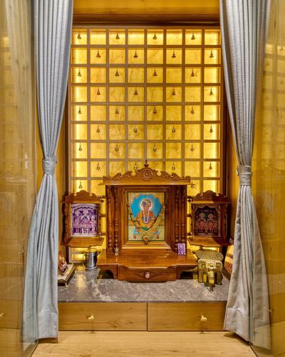 Backlit golden grid wall with hanging bells behind a carved wooden pooja unit, framed by grey curtains