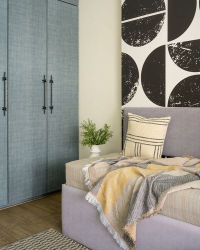Bedroom corner with grey upholstered bed, striped cushion, blue wardrobe and bold circular wallpaper.
