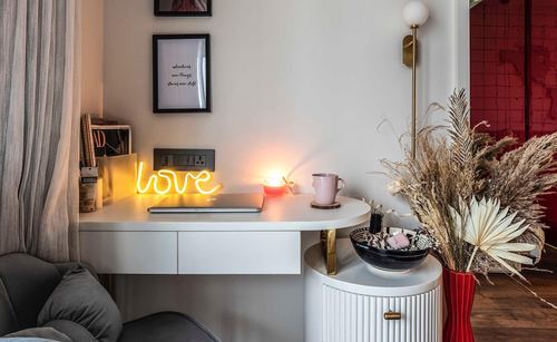 White curved study desk with neon love sign, framed prints, and dried floral arrangement beside red wall
