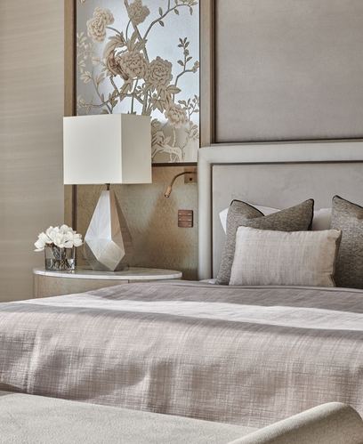 Neutral-toned bedroom corner with upholstered headboard, faceted lamp, and floral wall panel