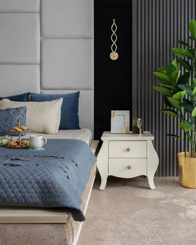 Bedroom corner with grey upholstered headboard, blue bedding, cream nightstand, and large plant against fluted wall