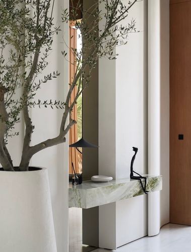 Curved white column beside marble console with sculpture, olive tree in planter, foyer
