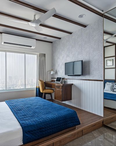 Bedroom with navy quilted bed on wooden platform, desk, TV, and city view window