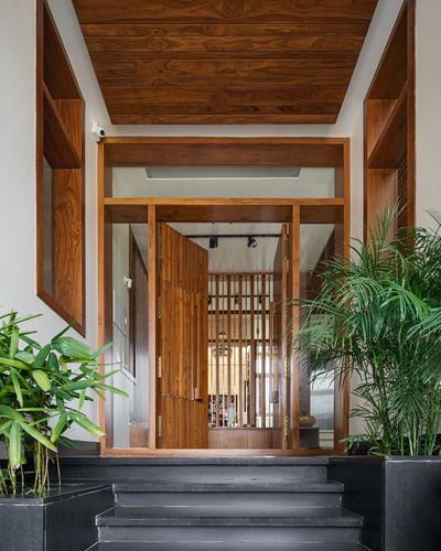Wood-framed entrance with pivot door, angled wood ceiling, and potted palms flanking dark stone steps