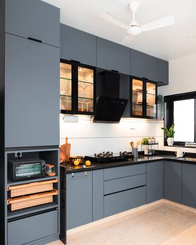L-shaped grey modular kitchen with black chimney hood, glass cabinets, and cane basket storage