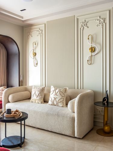 Beige living room with boucle sofa, ornate wall panels, brass sconces, and marble side tables