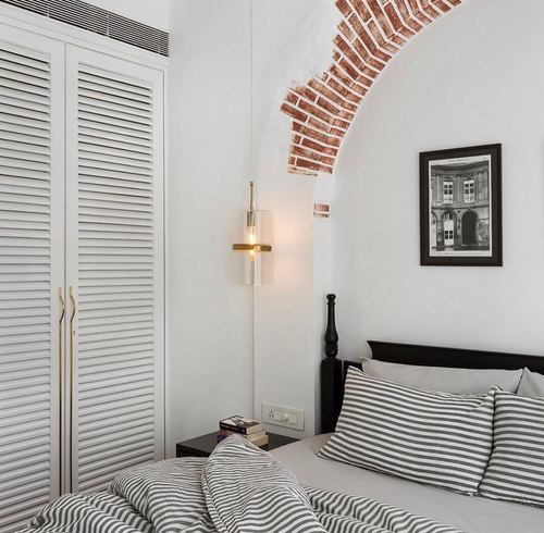 Colonial bedroom with brick arch, louvered wardrobe, brass sconce, and dark poster bed with striped linens