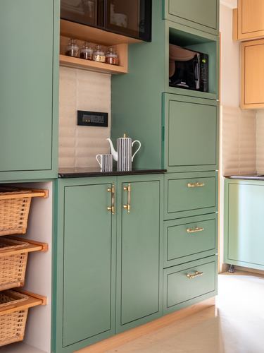 Sage green kitchen cabinetry with brass handles, open shelf, and cane pull-out baskets