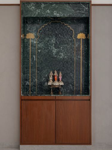 Green stone pooja niche with arched brass inlay above a wooden cabinet holding small idols