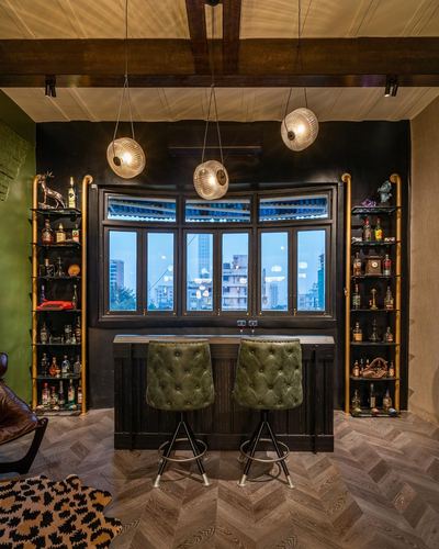 Home bar with green tufted stools, brass shelving, and bay window city view at dusk