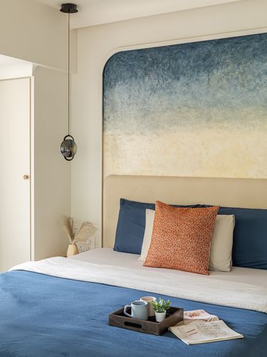 Bed with blue linens and terracotta cushion beneath a curved ombre-painted headboard and pendant light
