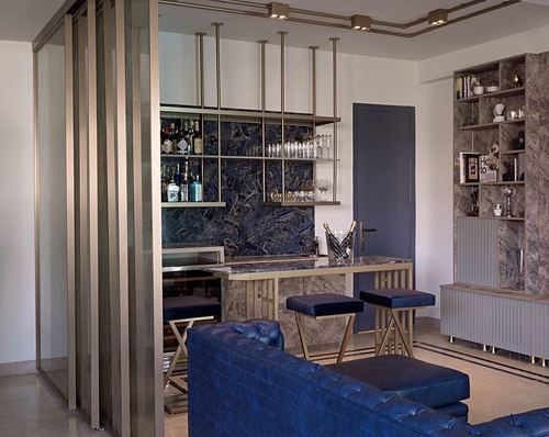 Home bar with dark marble backsplash, brass fittings, navy stools, and blue tufted sofa in living room
