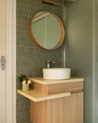 Round wood-framed mirror over vessel sink on tiered wooden vanity against scalloped green tile wall